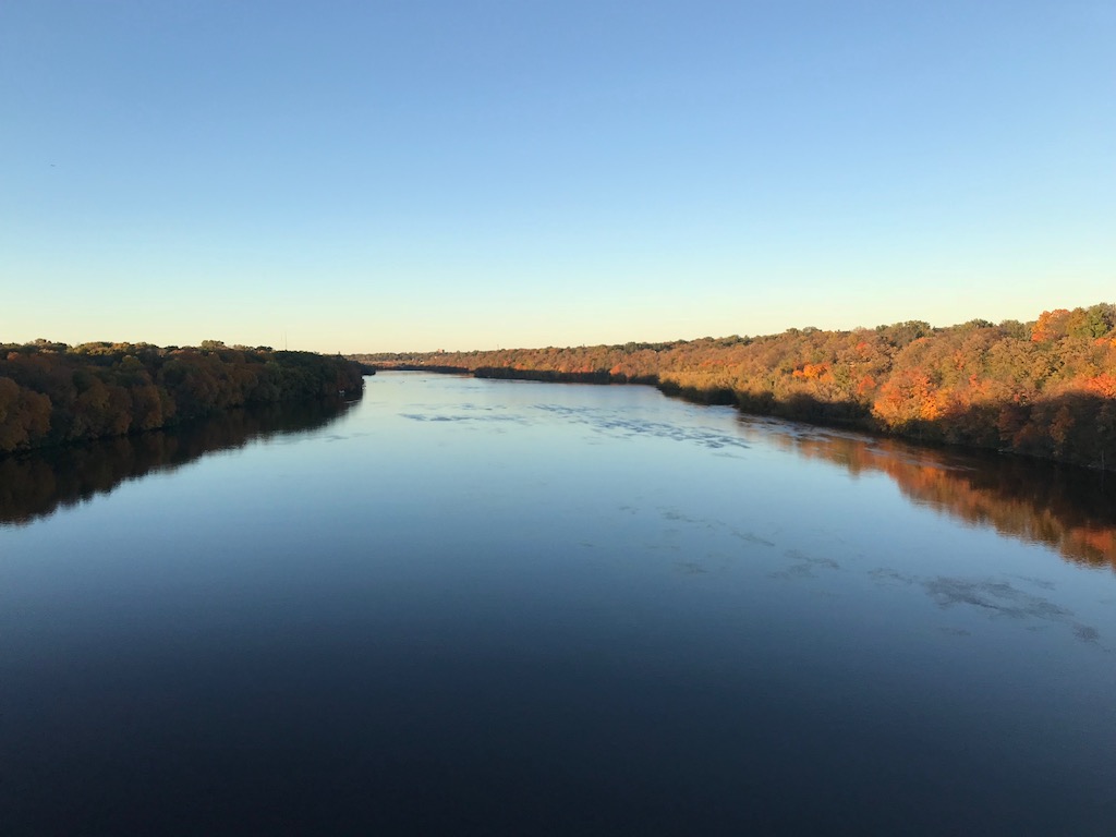 Photo of the Mississippi River at sunset. Fall colors are in full view along both banks. The Lake Street bridge is visible in the distance