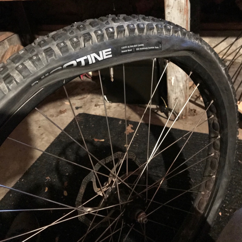 Photo of a mountain bike wheel, the tire features a prominent 'NICOTINE' logo and is almost, but not quite, installed onto the rim